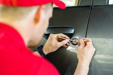 A male locksmith with a red shirt and hat servicing a car door lock of a dark silver car